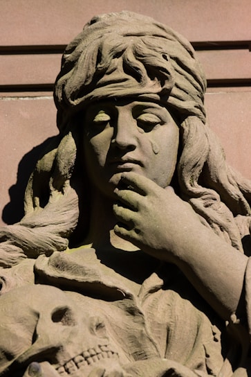A detailed stone sculpture of a woman with a sorrowful expression, featuring a tear on her cheek and a contemplative pose with her hand on her chin. The figure is adorned with a headpiece, and below the woman's face, there is a carving of a skull, adding a macabre element.