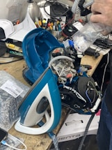 A disassembled steam iron is placed on a cluttered workbench. Electronic components and wires are visible within the open casing. Surrounding the iron are various tools, papers, and electronic parts. A person's hand is visible holding a plastic bag near the iron.