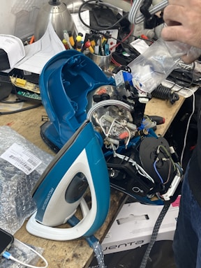 A disassembled steam iron is placed on a cluttered workbench. Electronic components and wires are visible within the open casing. Surrounding the iron are various tools, papers, and electronic parts. A person's hand is visible holding a plastic bag near the iron.