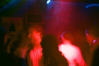 A dimly lit nightclub scene with vibrant red and blue lighting casting a dynamic atmosphere. Blurred figures suggest movement and dancing, adding to the sense of energy and activity.