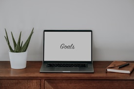 A wooden desk with a laptop displaying the word 'Goals' on its screen. To the left is a potted aloe vera plant, and to the right is a closed notebook with a pen on top.