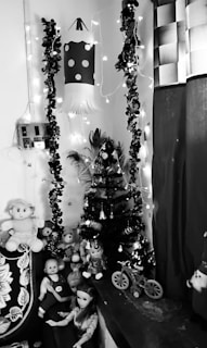 A small artificial Christmas tree is decorated with lights and ornaments, placed in the corner of a room. Surrounding the tree are various dolls and plush toys, giving the setting a playful and festive atmosphere. A paper lantern with a polka dot design hangs above, adding to the decorations. Strings of lights and garlands are draped along the walls, enhancing the scene's festive feel.