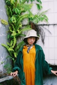 A person stands against a tiled wall covered with lush green ferns, wearing an oversized green corduroy coat, a mustard yellow dress, and a bucket hat with a blue scarf tied around their neck. The background features a mix of stone and vegetation, creating a natural and urban fusion.