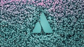 A geometric logo with two triangular shapes is embedded in a backdrop of numerous small spherical objects that transition from teal at the bottom to pink at the top.