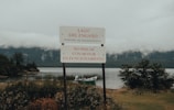 A sign stands prominently by the edge of a serene lake with a mountainous backdrop and low clouds. The sign reads 'Lago del Engaño' and warns against fishing with a motor in operation. The water is calm, with a small boat visible near the shoreline surrounded by lush greenery.