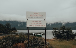 A sign stands prominently by the edge of a serene lake with a mountainous backdrop and low clouds. The sign reads 'Lago del Engaño' and warns against fishing with a motor in operation. The water is calm, with a small boat visible near the shoreline surrounded by lush greenery.