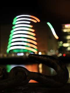 A blurred image of a spiral-shaped building adorned with bright lights in the colors of green, white, and orange in the background. In the foreground, a large, dark metal hook or chain link is partially visible, with reflections visible on a water surface.