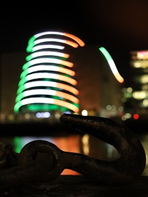 A blurred image of a spiral-shaped building adorned with bright lights in the colors of green, white, and orange in the background. In the foreground, a large, dark metal hook or chain link is partially visible, with reflections visible on a water surface.