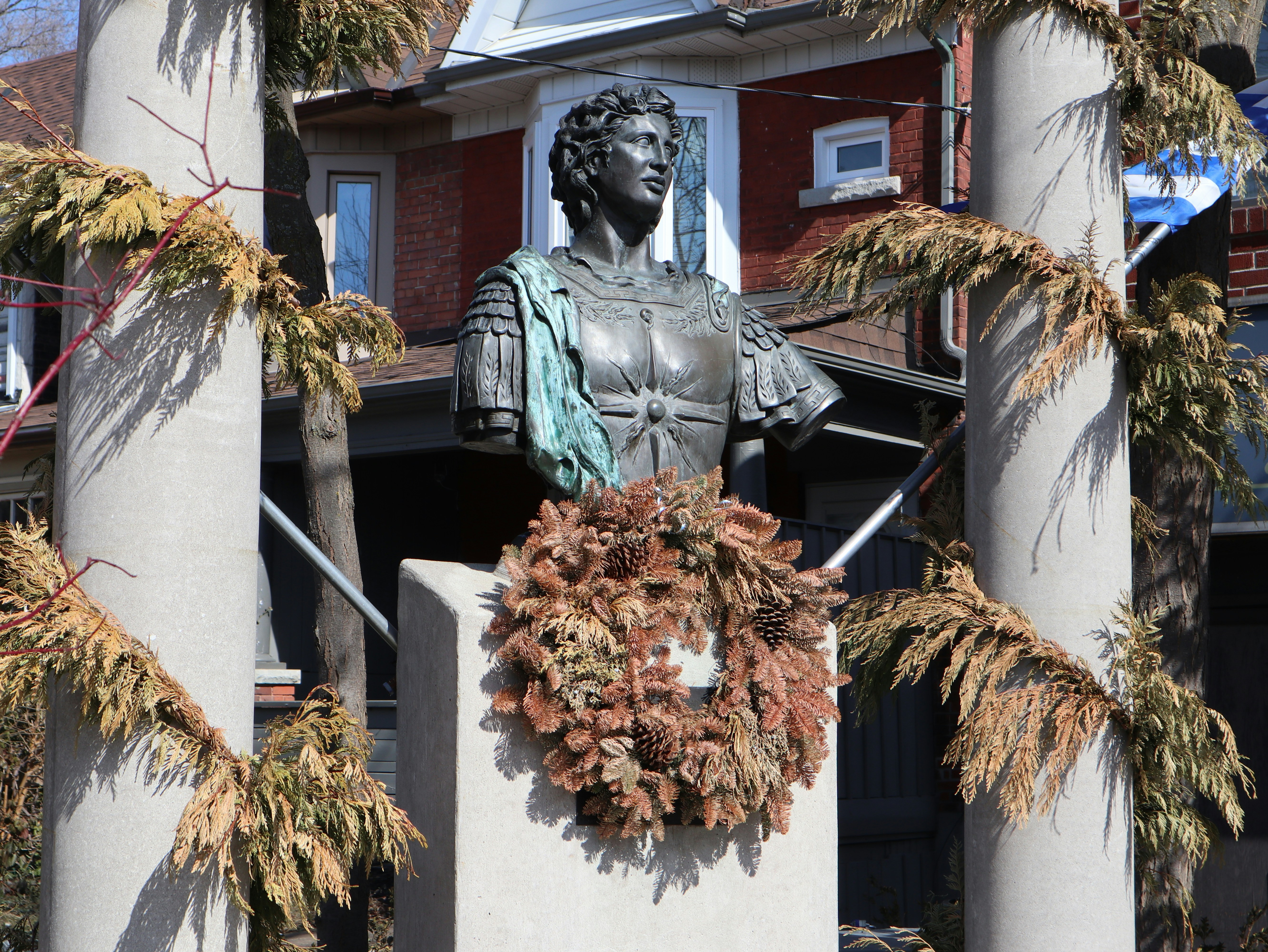a statue of a woman with a wreath around her neck, A bronze bust of Alexander The Great sits on a stage situated at the back of Alexander The Great Parkette in Toronto