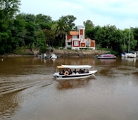 A small motorboat with passengers is moving along a brown river. In the background, a charming, multi-colored house with orange and white detailing stands surrounded by lush green trees and palm trees. Several other boats are docked along the riverbank.