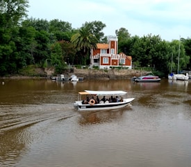 A small motorboat with passengers is moving along a brown river. In the background, a charming, multi-colored house with orange and white detailing stands surrounded by lush green trees and palm trees. Several other boats are docked along the riverbank.