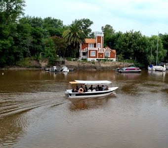 A small motorboat with passengers is moving along a brown river. In the background, a charming, multi-colored house with orange and white detailing stands surrounded by lush green trees and palm trees. Several other boats are docked along the riverbank.