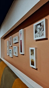 A series of framed pictures are arranged on a peach-colored wall. The frames vary in size and shape, displaying both photographs and artwork. One prominent picture features a person holding a camera. The setting includes a cushioned seating area with shades of brown and yellow.