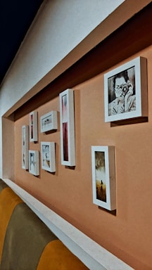 A series of framed pictures are arranged on a peach-colored wall. The frames vary in size and shape, displaying both photographs and artwork. One prominent picture features a person holding a camera. The setting includes a cushioned seating area with shades of brown and yellow.