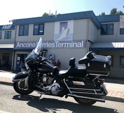 A black motorcycle is parked in front of a building with signage that reads 'Ancona Ferries Terminal'. The sleek design of the motorcycle is complemented by chrome details and a large windshield. The building in the background is gray with blue trim, and a logo featuring a silhouette of a ferry is visible. Several people are standing near the entrance of the building.