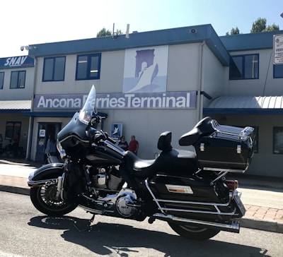 A black motorcycle is parked in front of a building with signage that reads 'Ancona Ferries Terminal'. The sleek design of the motorcycle is complemented by chrome details and a large windshield. The building in the background is gray with blue trim, and a logo featuring a silhouette of a ferry is visible. Several people are standing near the entrance of the building.