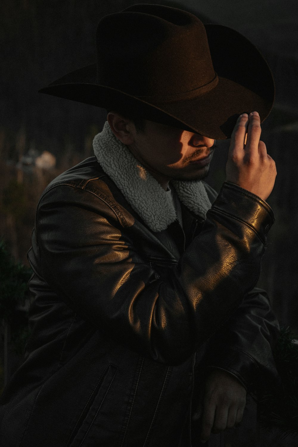 a man in a leather jacket and hat smoking a cigarette