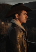 A person wearing a cowboy hat and leather jacket with a fur collar stands outdoors in a rustic setting, looking off into the distance. The background features blurred trees and hills, suggesting a mountainous or rural area bathed in warm, soft lighting.