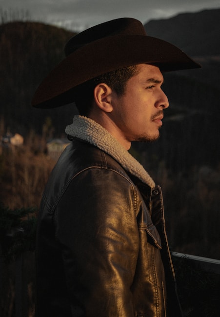A person wearing a cowboy hat and leather jacket with a fur collar stands outdoors in a rustic setting, looking off into the distance. The background features blurred trees and hills, suggesting a mountainous or rural area bathed in warm, soft lighting.