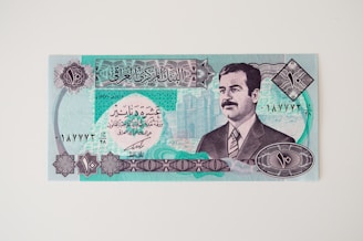An Iraqi banknote featuring intricate patterns and Arabic script, with the portrait of a man on the right side. The design includes geometric shapes and a detailed architectural illustration in the background.