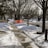 A residential street lined with bare trees on a winter day, with patches of snow on the ground and sidewalks. An orange construction barrier is placed across the sidewalk, and there are scattered residential homes in the background.