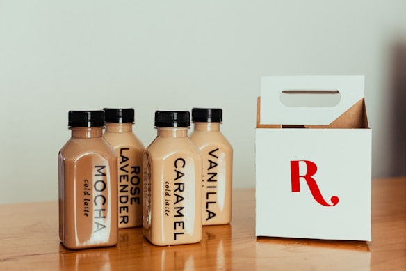 A group of four cold latte bottles with black caps is displayed on a wooden surface. Each bottle is labeled with different flavors: mocha, rose lavender, caramel, and vanilla. Next to the bottles is a white cardboard carrier with a large red letter 'R' on it.