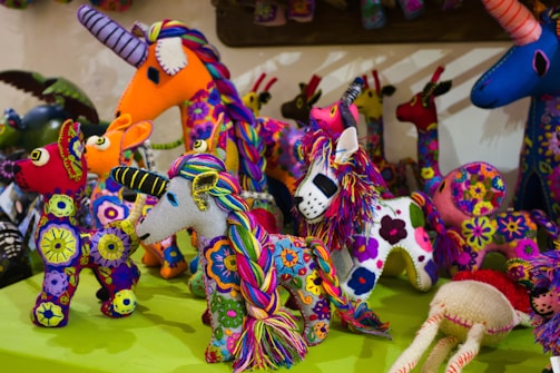 A collection of brightly colored, intricately embroidered stuffed animal toys with various designs including unicorns and llamas. Each toy features vibrant patterns and multicolored threads, creating a festive and artistic display.