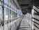 A concrete walkway with metal railings runs alongside a large wall of windows, casting angular shadows across the floor from the sunlight streaming in. The architectural style features a modern, industrial aesthetic, with visible support beams and a combination of metal and glass elements.