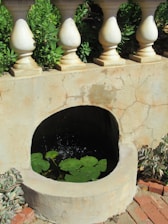 A stone wall with rounded pillars on top, behind which lush green plants can be seen. A small, circular pond embedded within the wall contains lily pads and reflects light with gentle water ripples.