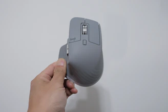 A hand is holding a gray ergonomic computer mouse with a vertical orientation. The mouse has a scroll wheel and several buttons, highlighted in a minimalist design. The background is a plain, light-colored wall.