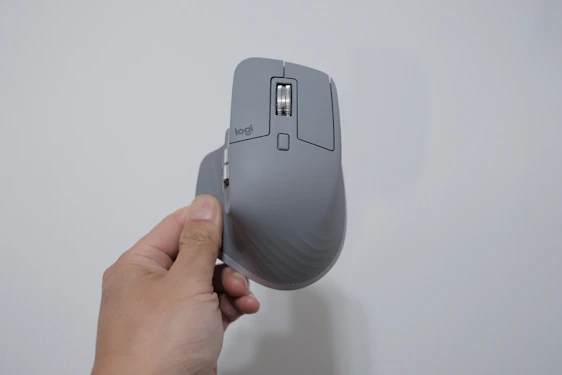 A hand is holding a gray ergonomic computer mouse with a vertical orientation. The mouse has a scroll wheel and several buttons, highlighted in a minimalist design. The background is a plain, light-colored wall.