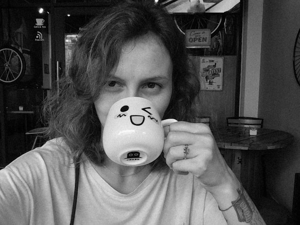 A smiling young woman holding a manga volume, sitting in a cafe decorated with anime art.