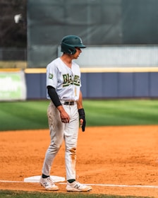 A baseball player stands on a baseball field wearing a uniform with the word 'Blazers' and the number 1. The athlete wears a green helmet and appears to be on a base, with one foot on the bag. The field has a grass outfield and a dirt infield.