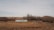 A solitary mobile home is situated in a barren, flat landscape surrounded by leafless trees and dry, brown grass. Two old vehicles are parked nearby, and the horizon stretches out to reveal rolling hills and a cloudy sky.