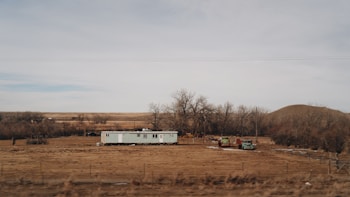 A solitary mobile home is situated in a barren, flat landscape surrounded by leafless trees and dry, brown grass. Two old vehicles are parked nearby, and the horizon stretches out to reveal rolling hills and a cloudy sky.