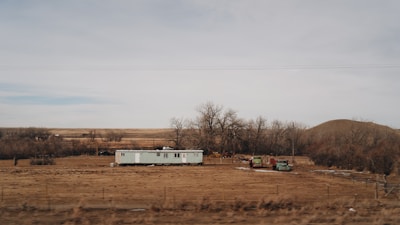 A solitary mobile home is situated in a barren, flat landscape surrounded by leafless trees and dry, brown grass. Two old vehicles are parked nearby, and the horizon stretches out to reveal rolling hills and a cloudy sky.
