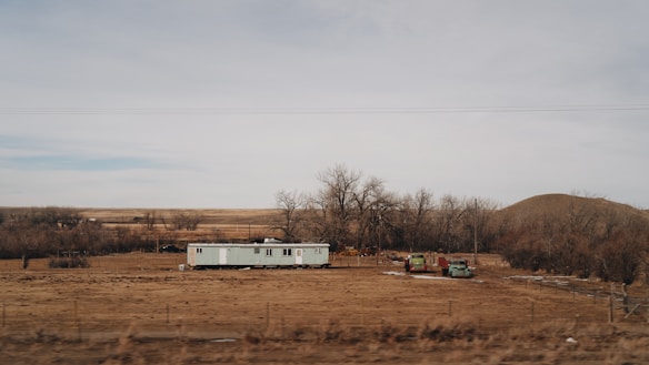 A solitary mobile home is situated in a barren, flat landscape surrounded by leafless trees and dry, brown grass. Two old vehicles are parked nearby, and the horizon stretches out to reveal rolling hills and a cloudy sky.