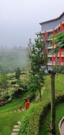 A lush green landscape with a multi-story building on the right side. The building has gray walls with orange accents, likely balconies or windows. The foreground features a lawn with neatly trimmed bushes and trees. Potted plants add variety to the garden scenery. A red fire hydrant and a matching red object are visible near a stone walkway that curves through the grass.