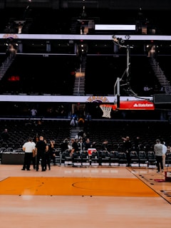 An indoor basketball court with empty seating areas, a basketball hoop positioned centrally, and a few people gathered near the sideline. The arena appears to be dimly lit with a focus on the court.
