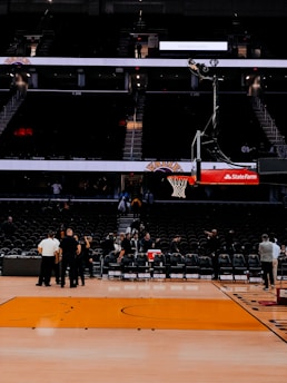 An indoor basketball court with empty seating areas, a basketball hoop positioned centrally, and a few people gathered near the sideline. The arena appears to be dimly lit with a focus on the court.
