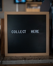 A wooden-framed black letter board with the words 'COLLECT HERE' written in white letters is placed on a countertop. The background is blurred and appears to be a cafe or restaurant setting.
