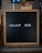 A wooden-framed black letter board with the words 'COLLECT HERE' written in white letters is placed on a countertop. The background is blurred and appears to be a cafe or restaurant setting.