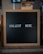 A wooden-framed black letter board with the words 'COLLECT HERE' written in white letters is placed on a countertop. The background is blurred and appears to be a cafe or restaurant setting.