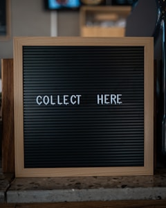 A wooden-framed black letter board with the words 'COLLECT HERE' written in white letters is placed on a countertop. The background is blurred and appears to be a cafe or restaurant setting.