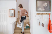 A woman carrying a cleaning caddy and broom up tiled stairs in a home