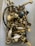 A collection of assorted metal hardware, including handles, knobs, and screws, placed loosely inside a plastic container. The objects are primarily made of brass with some white ceramic elements, and a few pieces are painted black or feature different metallic finishes.