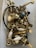 A collection of assorted metal hardware, including handles, knobs, and screws, placed loosely inside a plastic container. The objects are primarily made of brass with some white ceramic elements, and a few pieces are painted black or feature different metallic finishes.