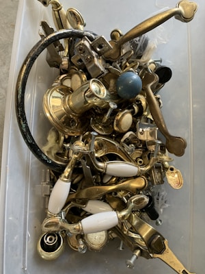 A collection of assorted metal hardware, including handles, knobs, and screws, placed loosely inside a plastic container. The objects are primarily made of brass with some white ceramic elements, and a few pieces are painted black or feature different metallic finishes.