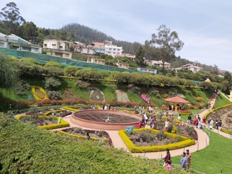 A well-maintained garden with vibrant flower beds arranged in intricate patterns. A circular path surrounds a sundial at the center. Visitors stroll along pathways, while a collection of shrubs and trees lines the background. Houses and buildings are visible on a gently sloping hill, with a dense forest in the far distance.