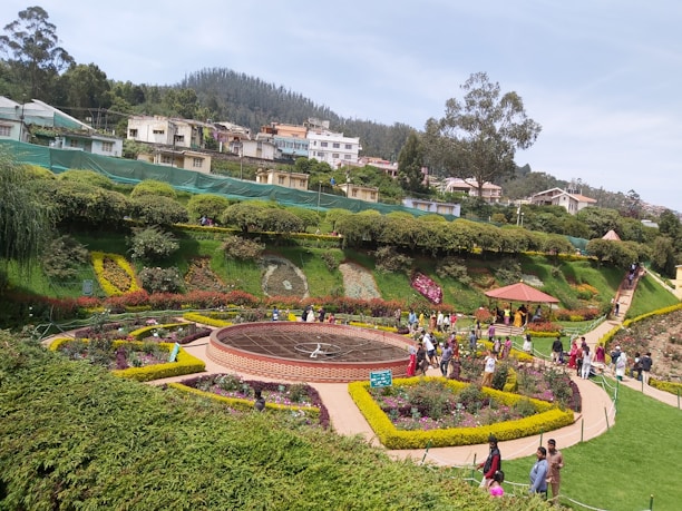A well-maintained garden with vibrant flower beds arranged in intricate patterns. A circular path surrounds a sundial at the center. Visitors stroll along pathways, while a collection of shrubs and trees lines the background. Houses and buildings are visible on a gently sloping hill, with a dense forest in the far distance.